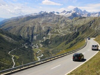 Wohnmobil auf kurvenreicher Alpenstraße vor verschneiten Berggipfeln